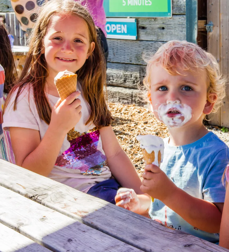 Three children eating ice creams in waffle cones at a farm park picnic table