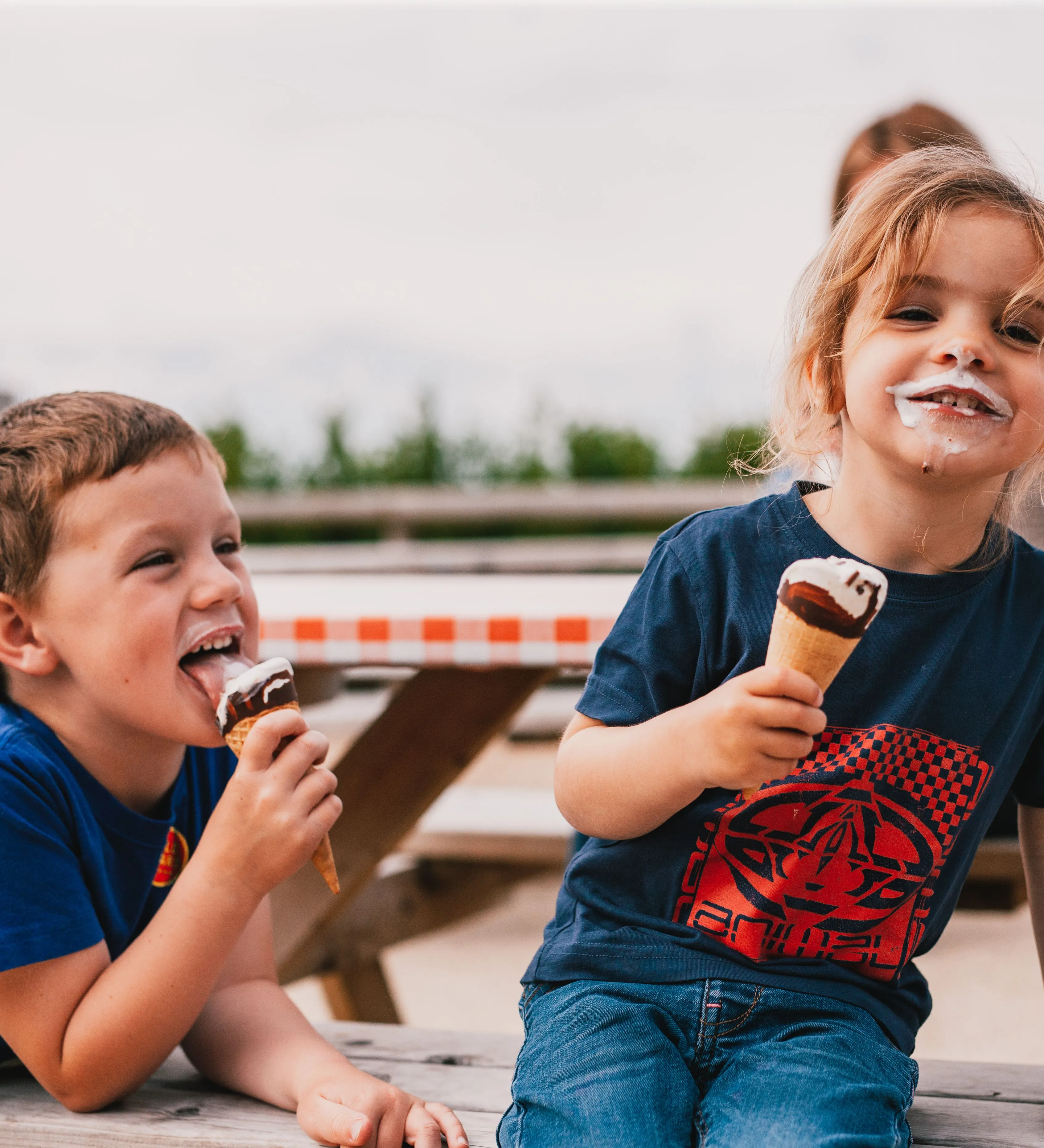 Two children eating ice cream cones at a picnic table, one smiling with ice cream on their face