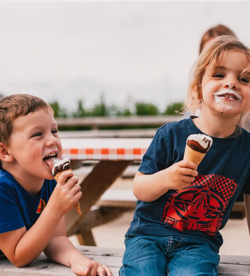 Two children eating ice cream cones at a picnic table, one smiling with ice cream on their face