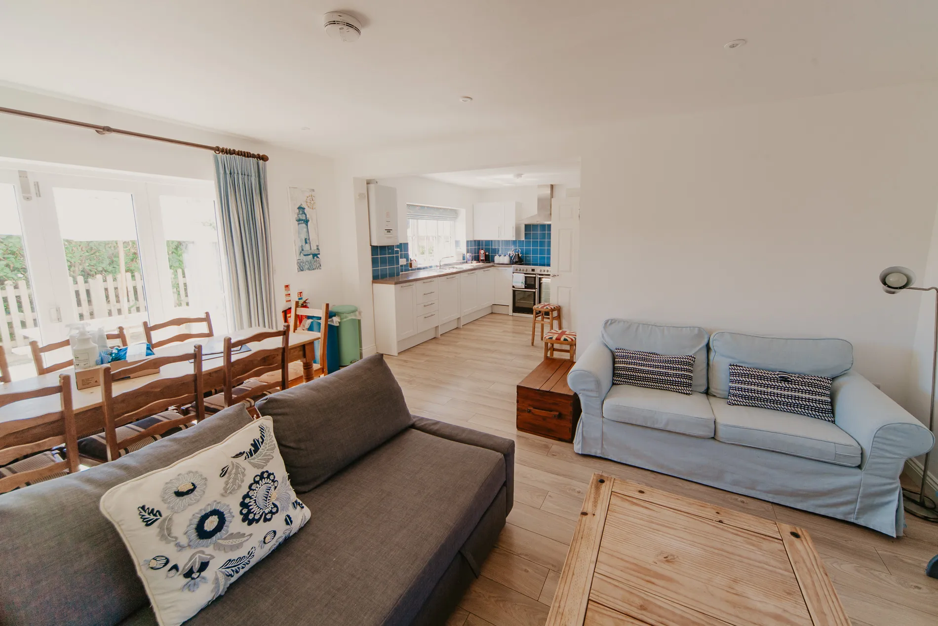 Open-plan cottage living and dining area with sofas, wooden table and white kitchen with blue tiled splashback
