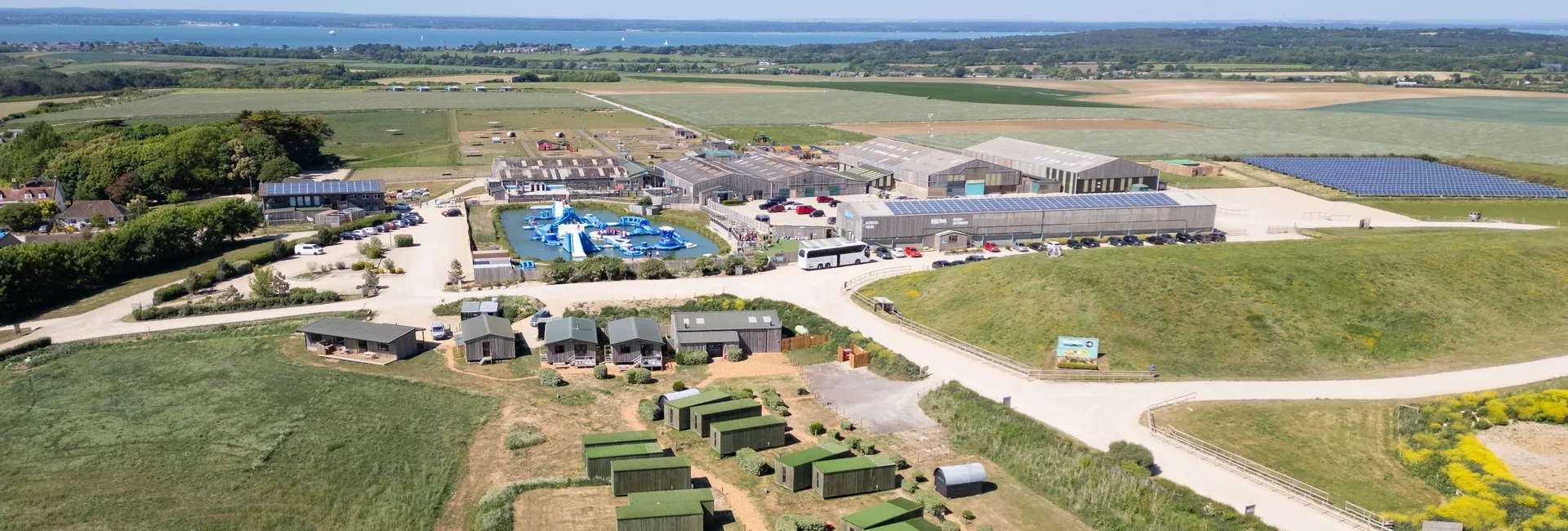 Aerial view of Tapnell Farm and Camp Tapnell with glamping cabins, farm buildings and an outdoor water park, with countryside and sea in the distance.