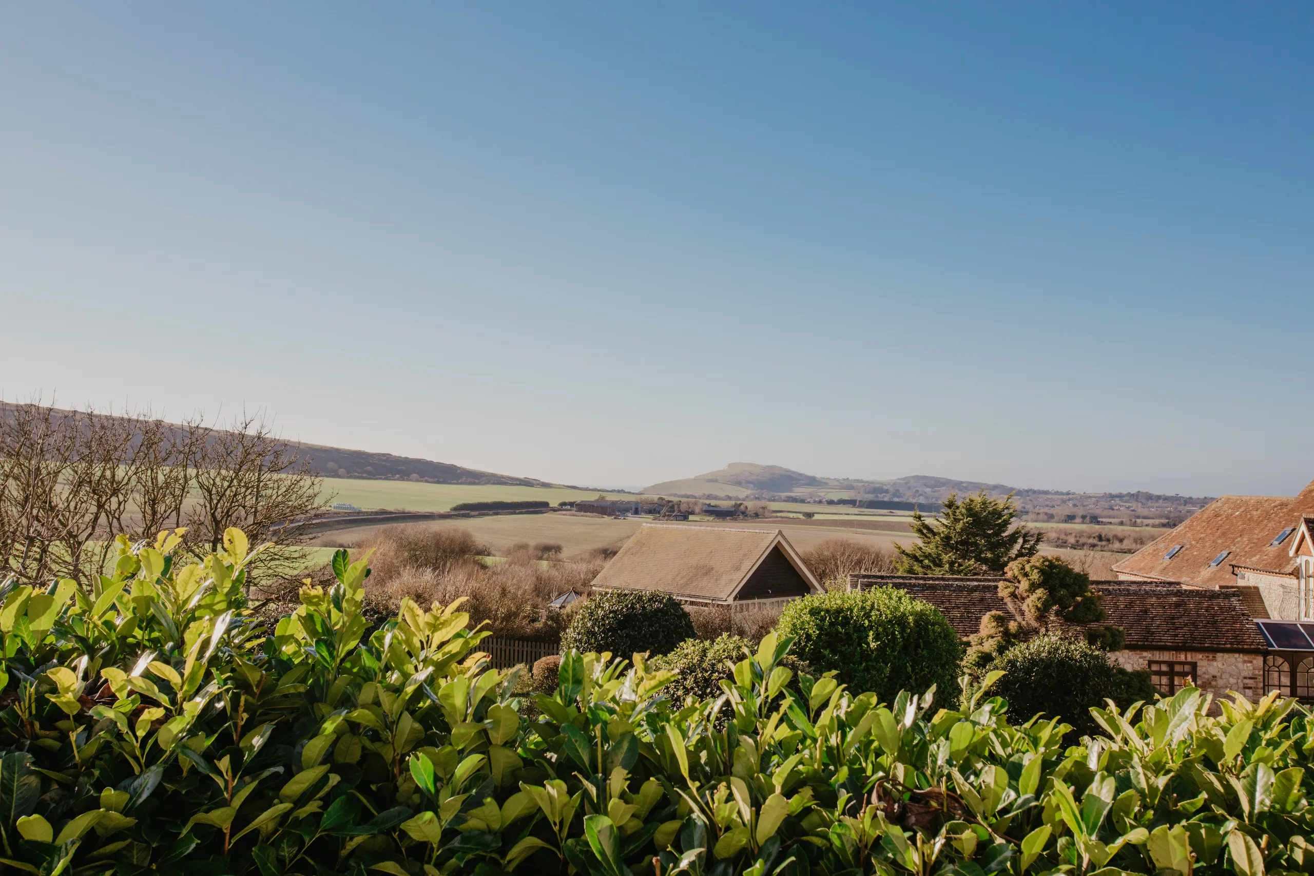 View over green hedgerow to cottage rooftops and rolling countryside under a clear blue sky