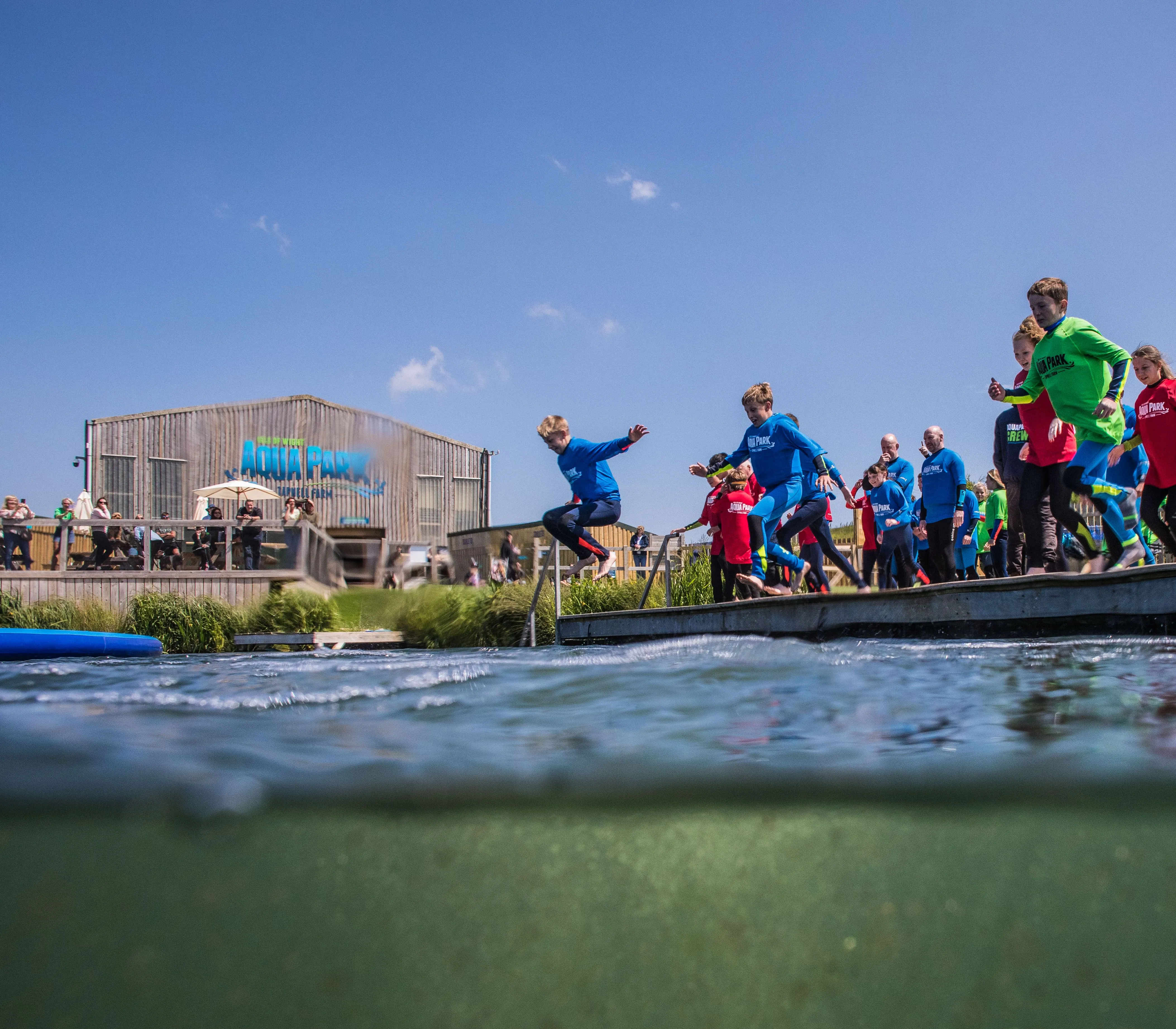 Children in colourful wetsuits jump from a platform into open water at an aqua park
