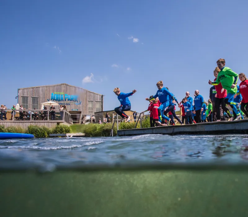 Children in colourful wetsuits jump from a platform into open water at an aqua park