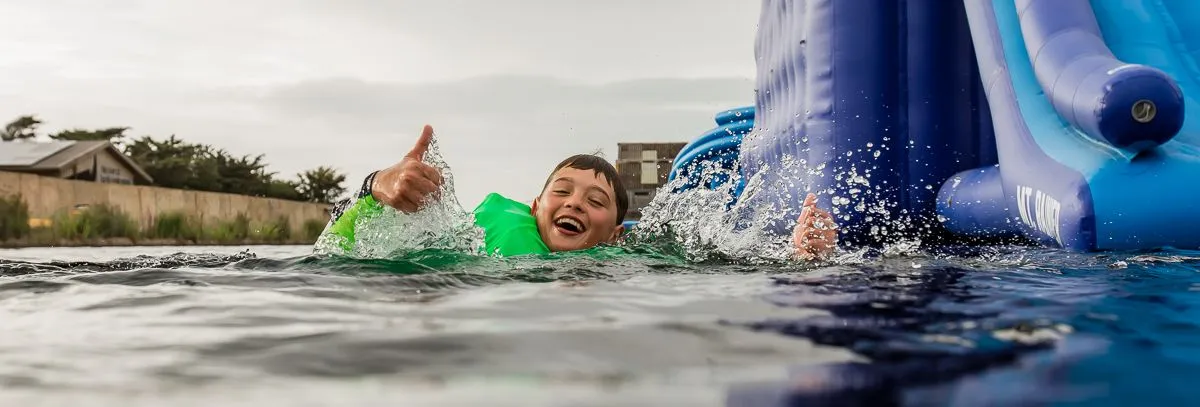 Child in a green buoyancy aid gives a thumbs up in the water beside a blue inflatable slide at the Isle of Wight Aqua Park