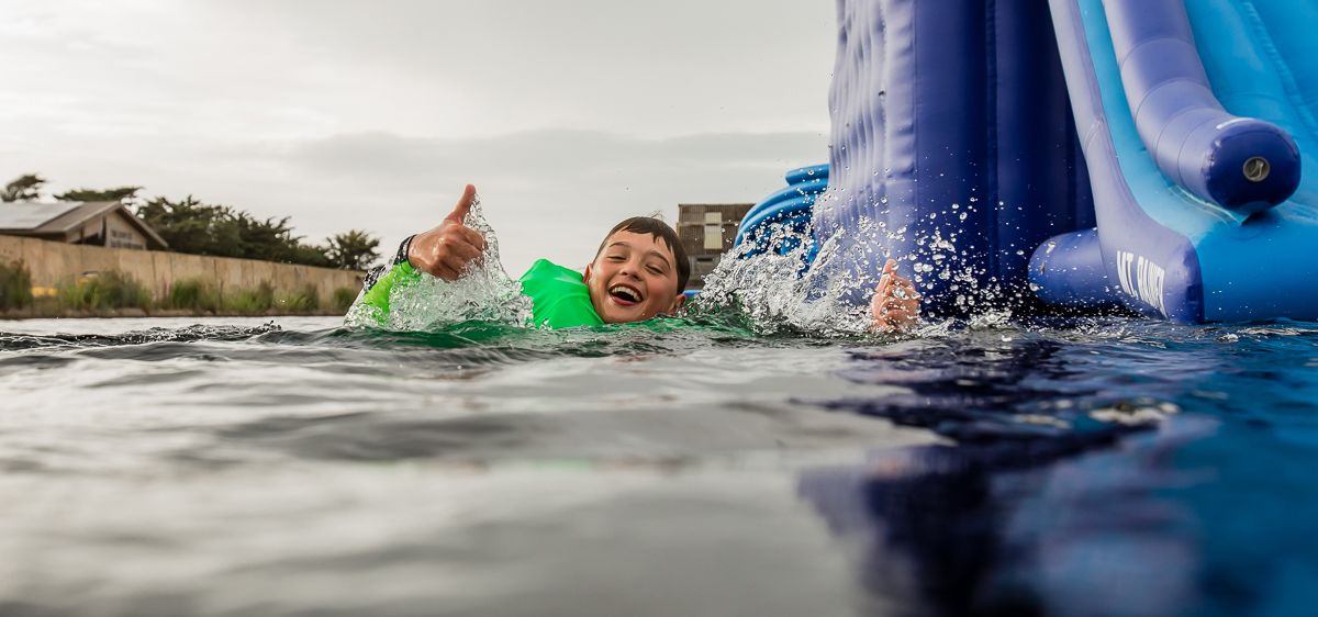 Child in a green buoyancy aid gives a thumbs up in the water beside a blue inflatable slide at the Isle of Wight Aqua Park