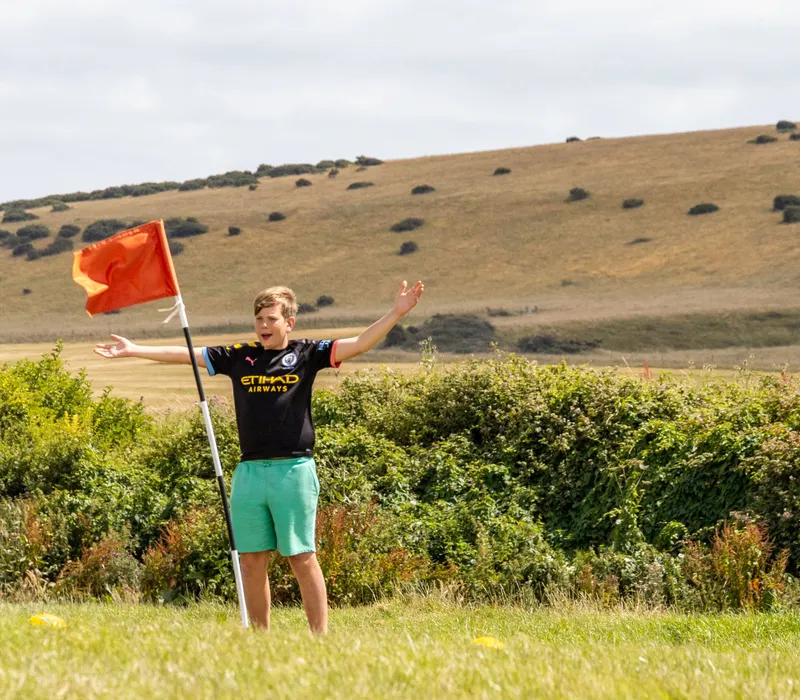 Boy standing beside a red flag on the football golf course at Tapnell Farm with rolling downs in the background