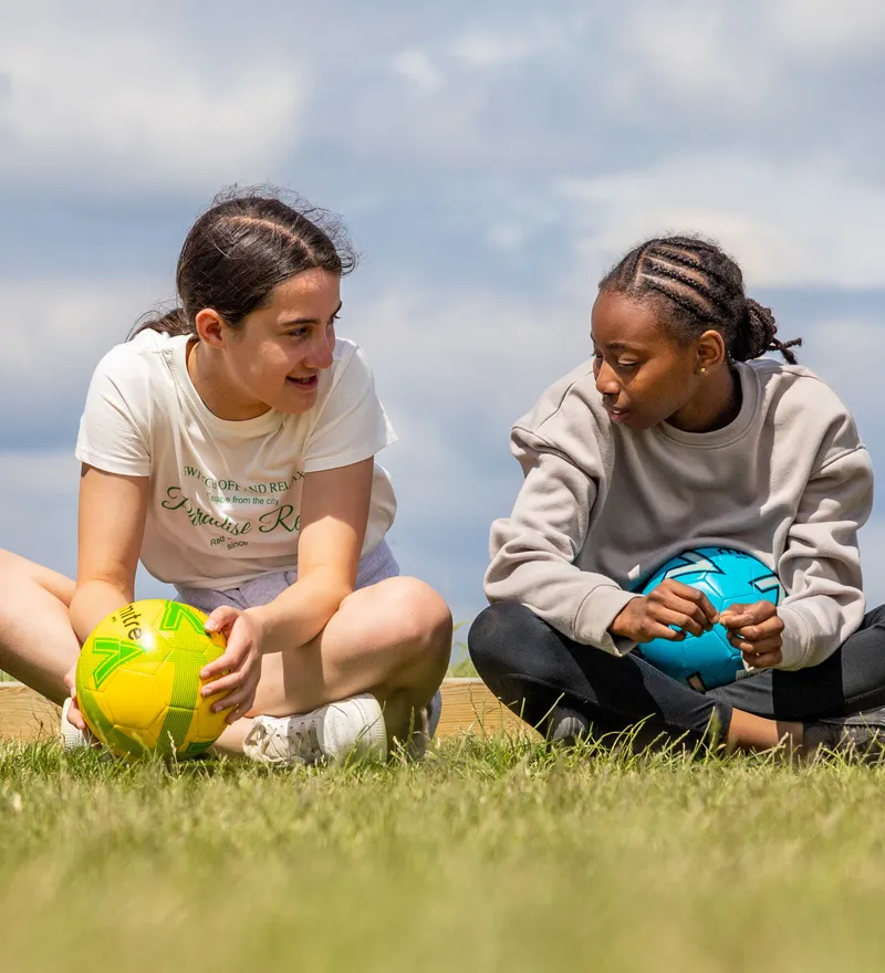 Three young people sitting on grass with colourful footballs, chatting outdoors