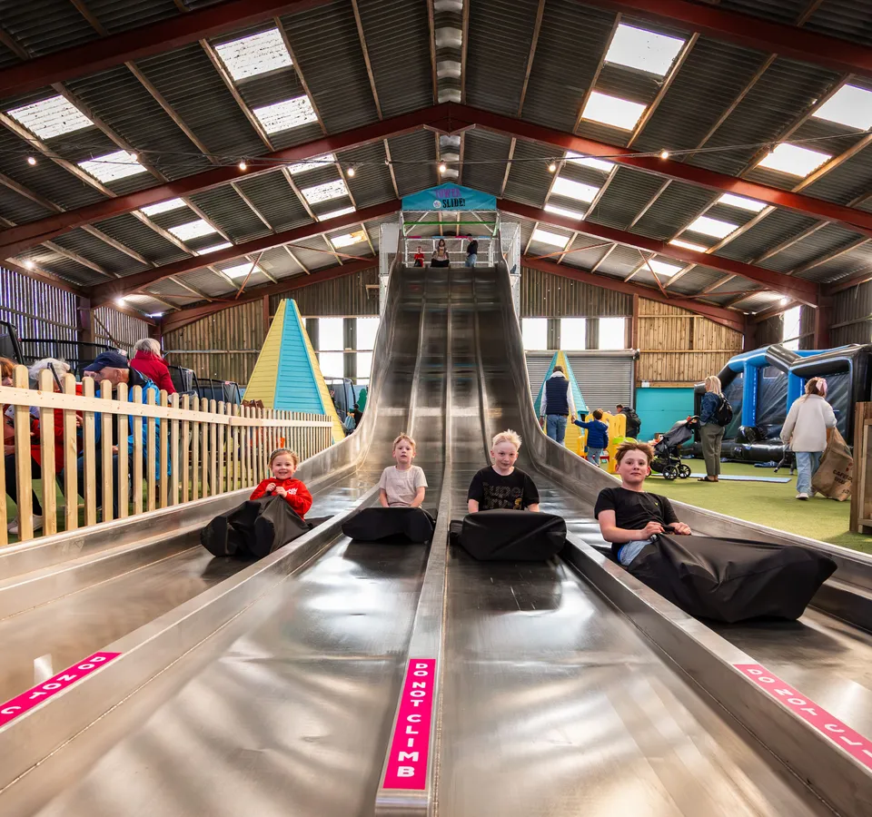 Children sitting on black sliding mats at the bottom of the Tower Slide in an indoor barn play area at Tapnell Farm Park