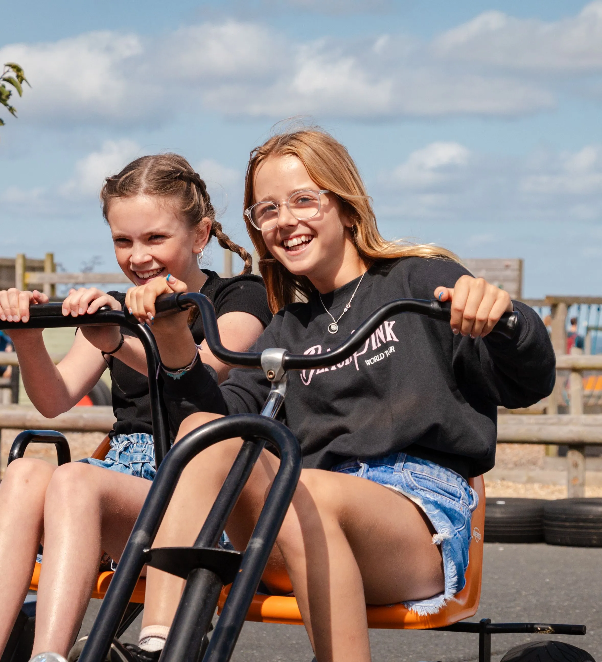Two girls smiling while riding a pedal go-kart at an outdoor farm park on a sunny day