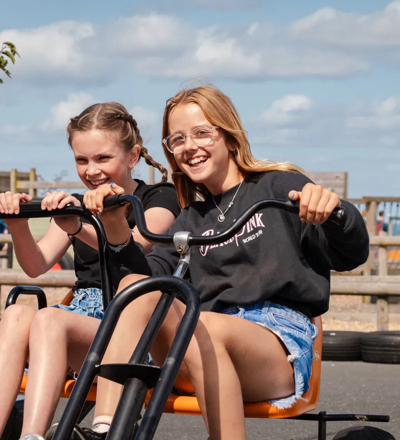 Two girls smiling while riding a pedal go-kart at an outdoor farm park on a sunny day