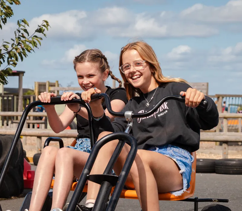 Two girls smiling while riding a pedal go-kart at an outdoor farm park on a sunny day