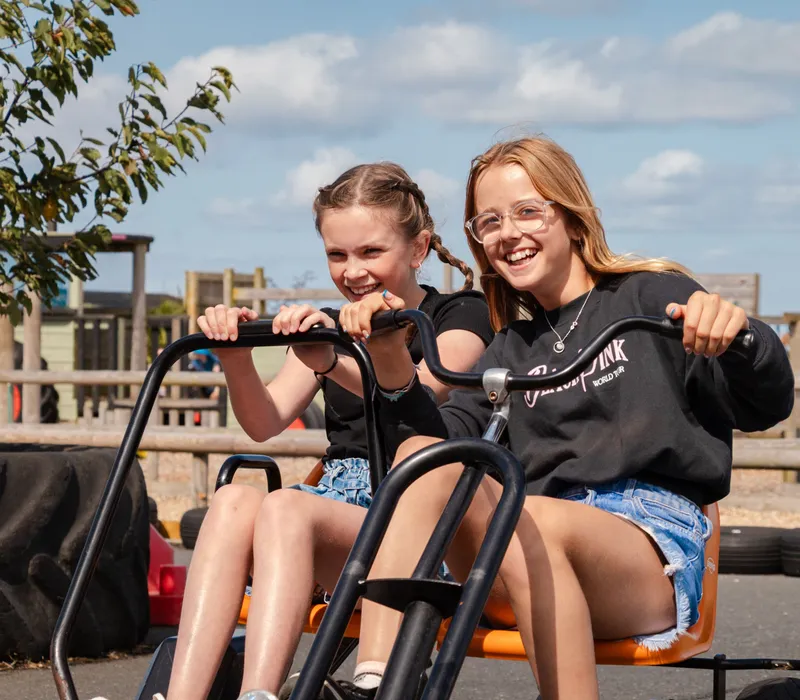Two girls smiling while riding a pedal go-kart at an outdoor farm park on a sunny day