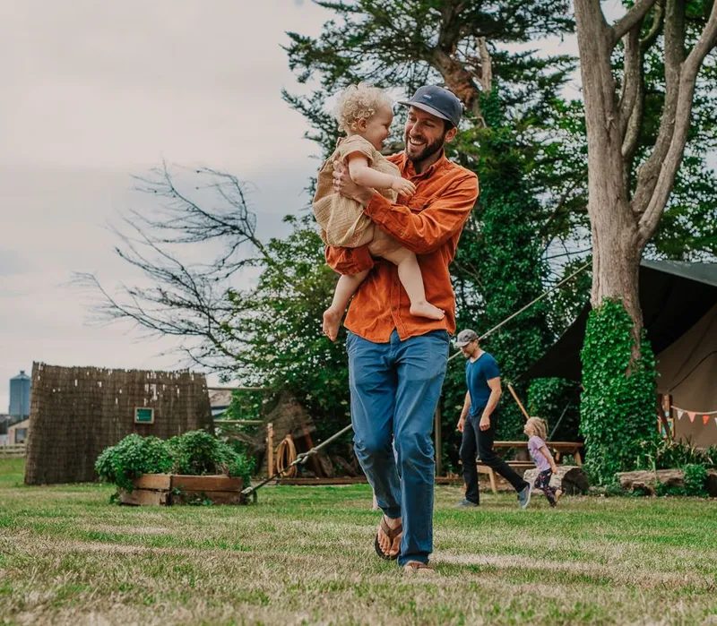 Man carrying a toddler and smiling in a grassy campsite near a large tent