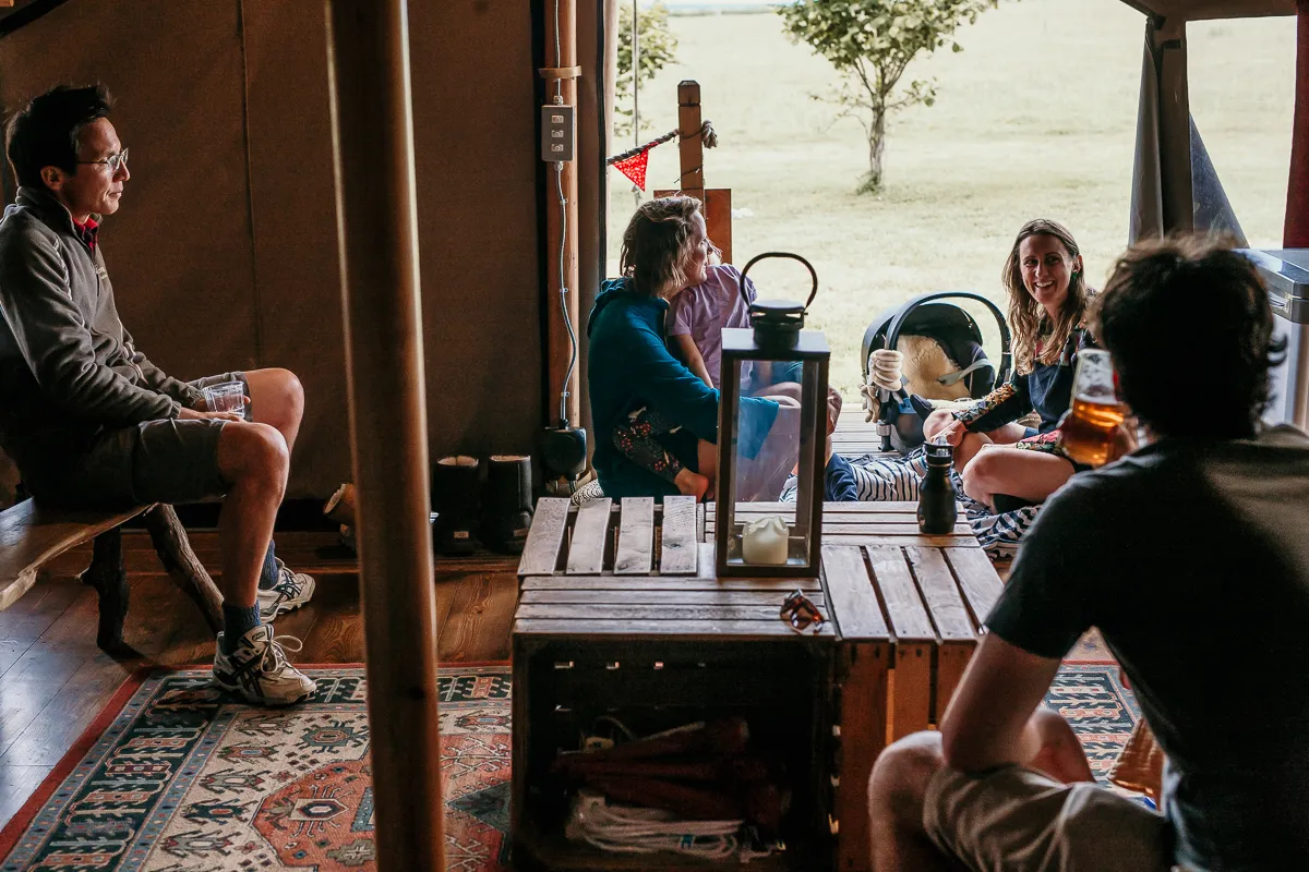 Group of friends relaxing inside a canvas tent with drinks around a wooden table, looking out onto a grassy field