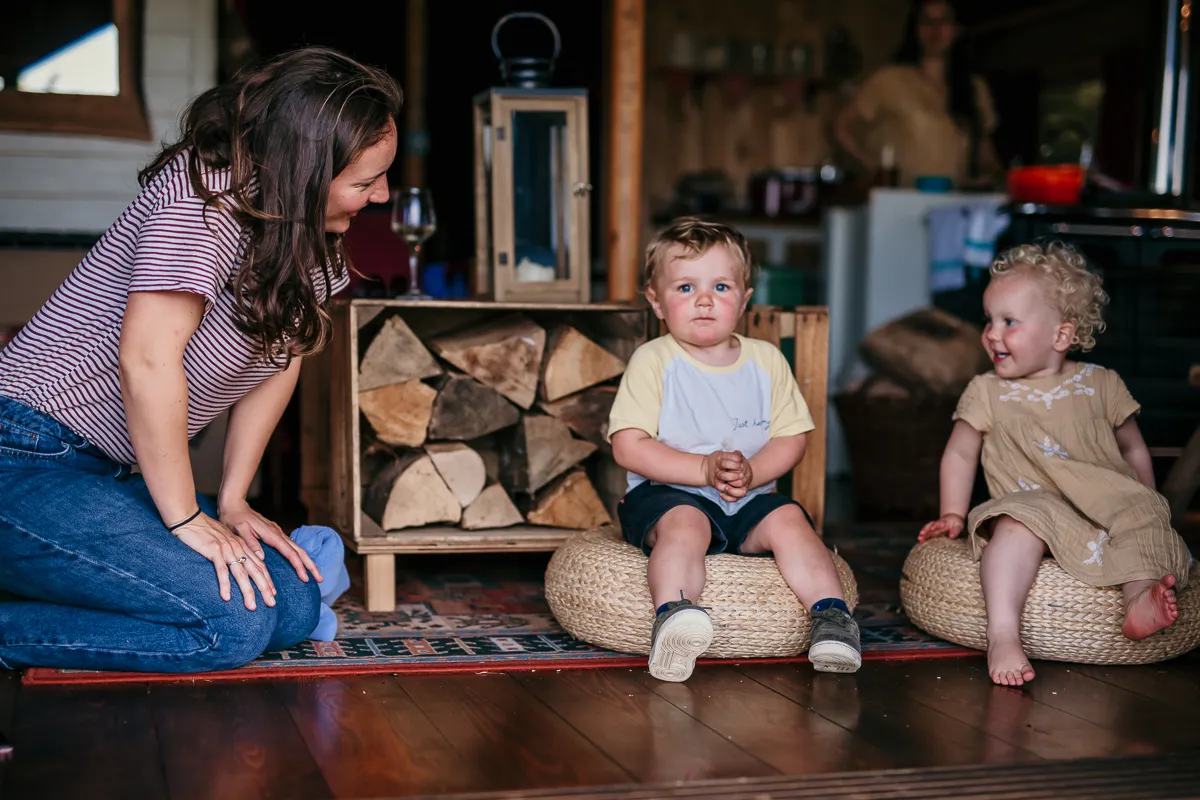 Woman kneeling beside two toddlers sitting on woven floor cushions in a rustic, cosy room with stacked firewood.