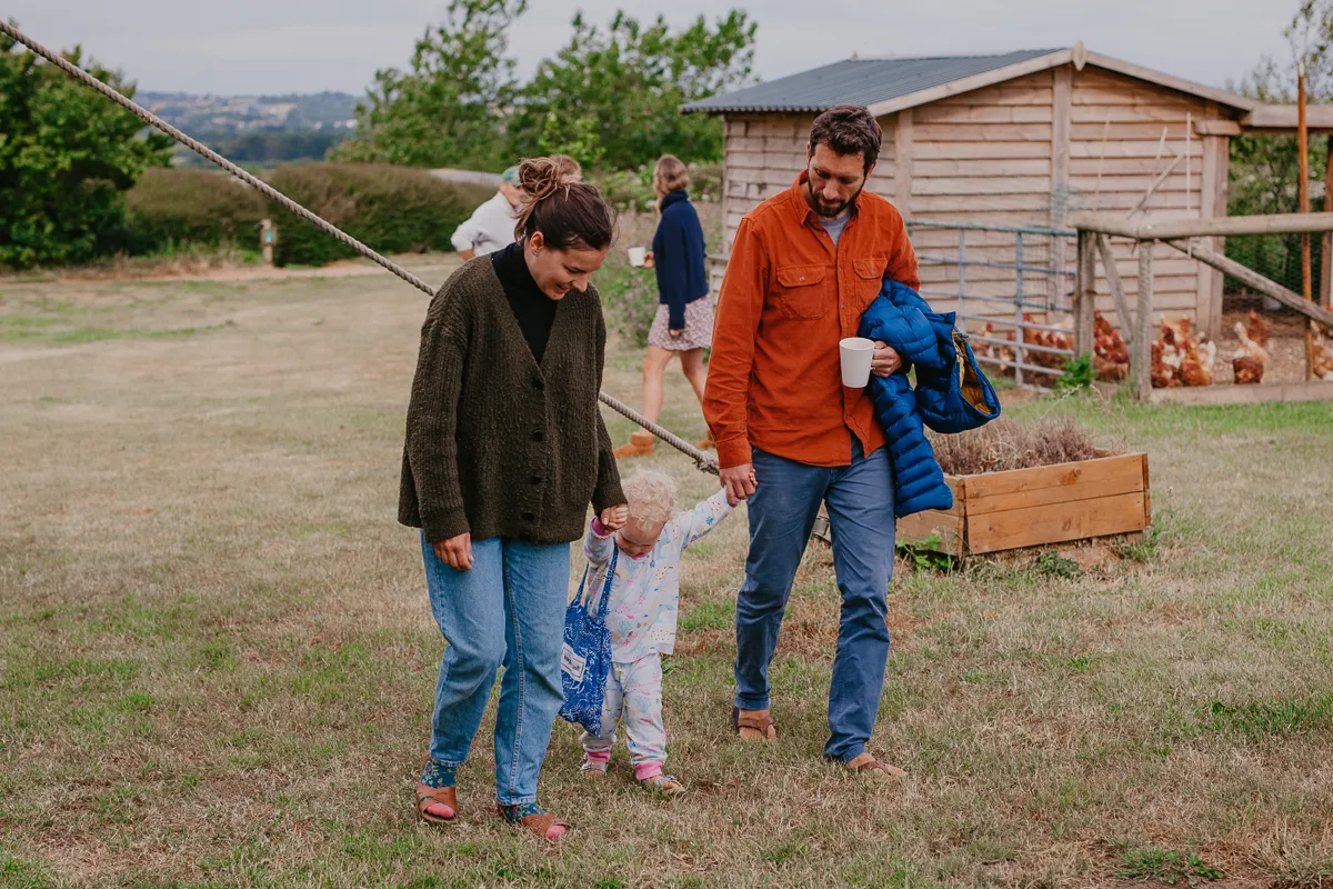 Family walking with a small child across a grassy farmyard near a chicken coop