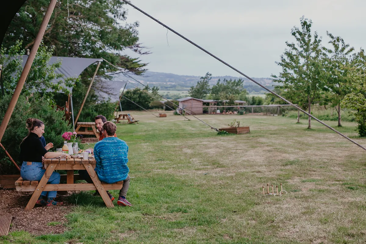Three people sitting at a picnic table outdoors in a grassy campsite with trees and hills in the background