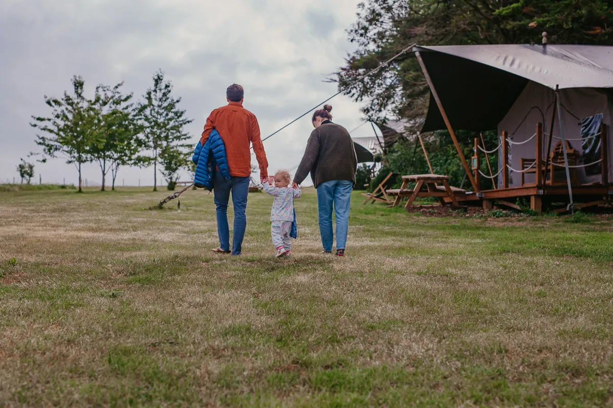 Parents holding a toddler’s hands as they walk across a grassy field towards safari tents at Tapnell Farm