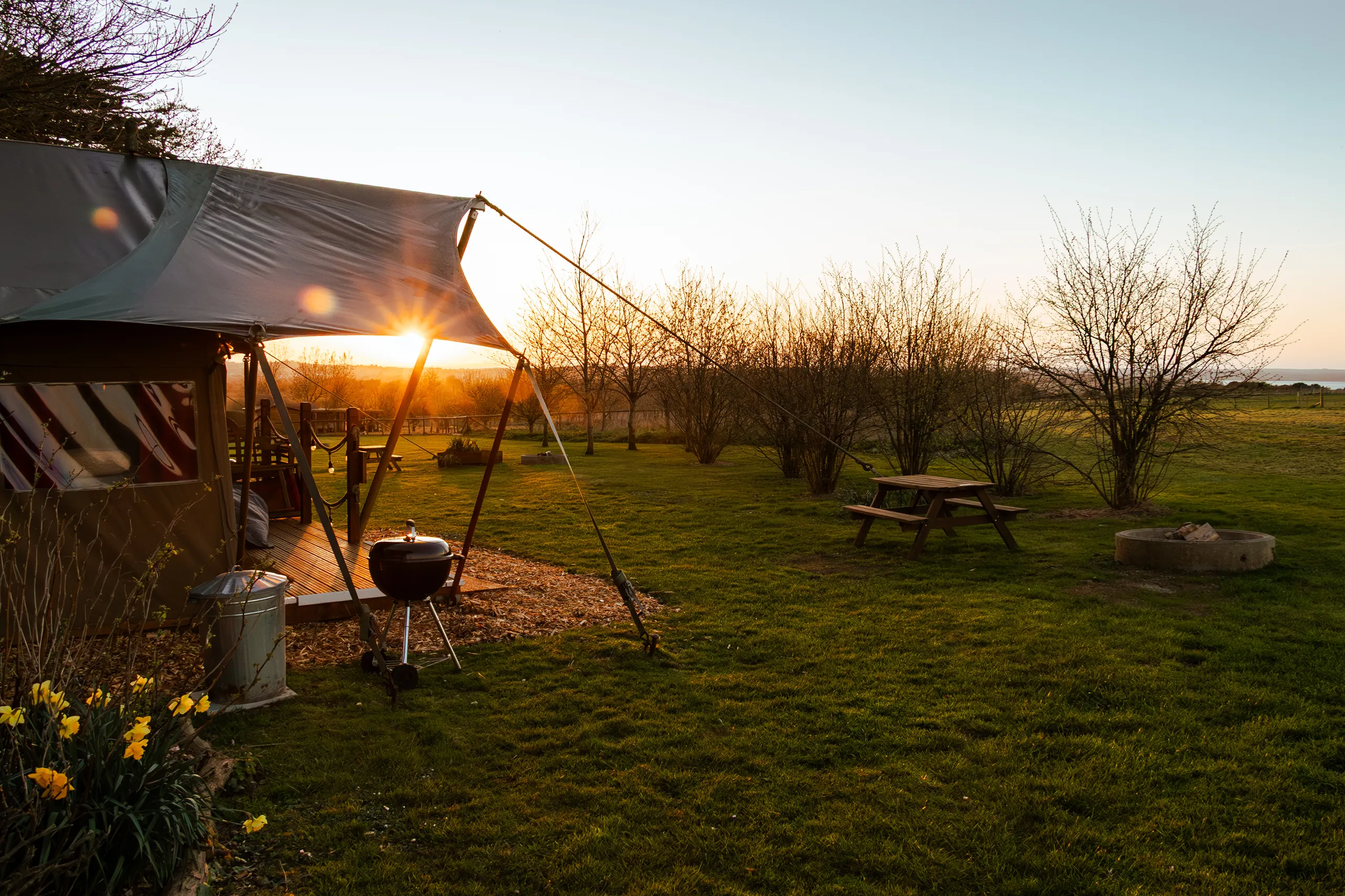 Sun setting behind a safari-style glamping tent with a picnic table and fire pit on a grassy field