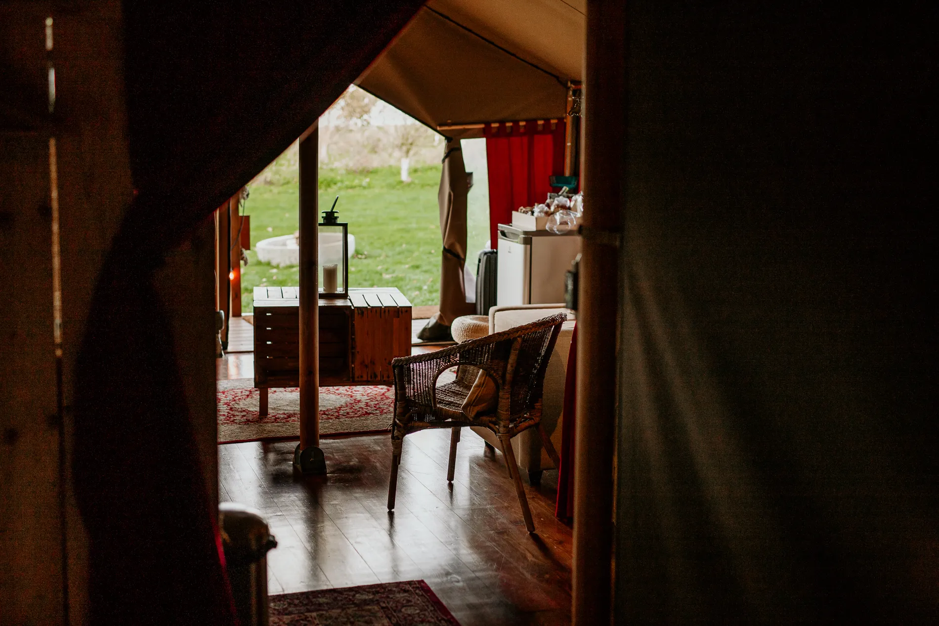 Cosy safari tent interior with wicker chair, wooden furniture and red curtains opening onto a grassy view