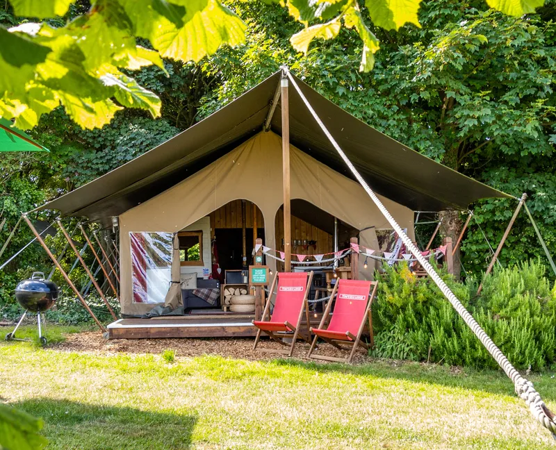 Safari-style eco lodge tent at Tapnell Farm on the Isle of Wight, with deck chairs and a barbecue in a leafy woodland setting