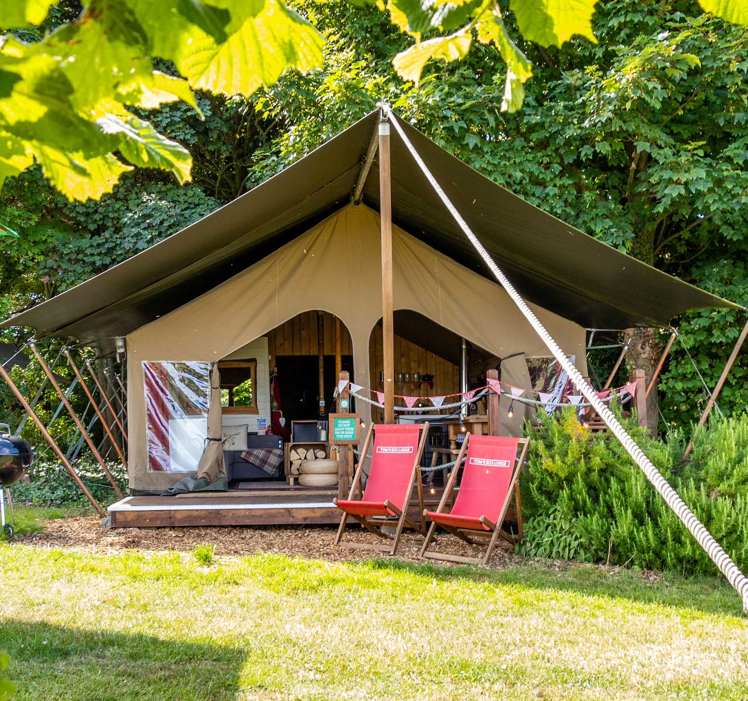 Safari-style eco lodge tent at Tapnell Farm on the Isle of Wight, with deck chairs and a barbecue in a leafy woodland setting
