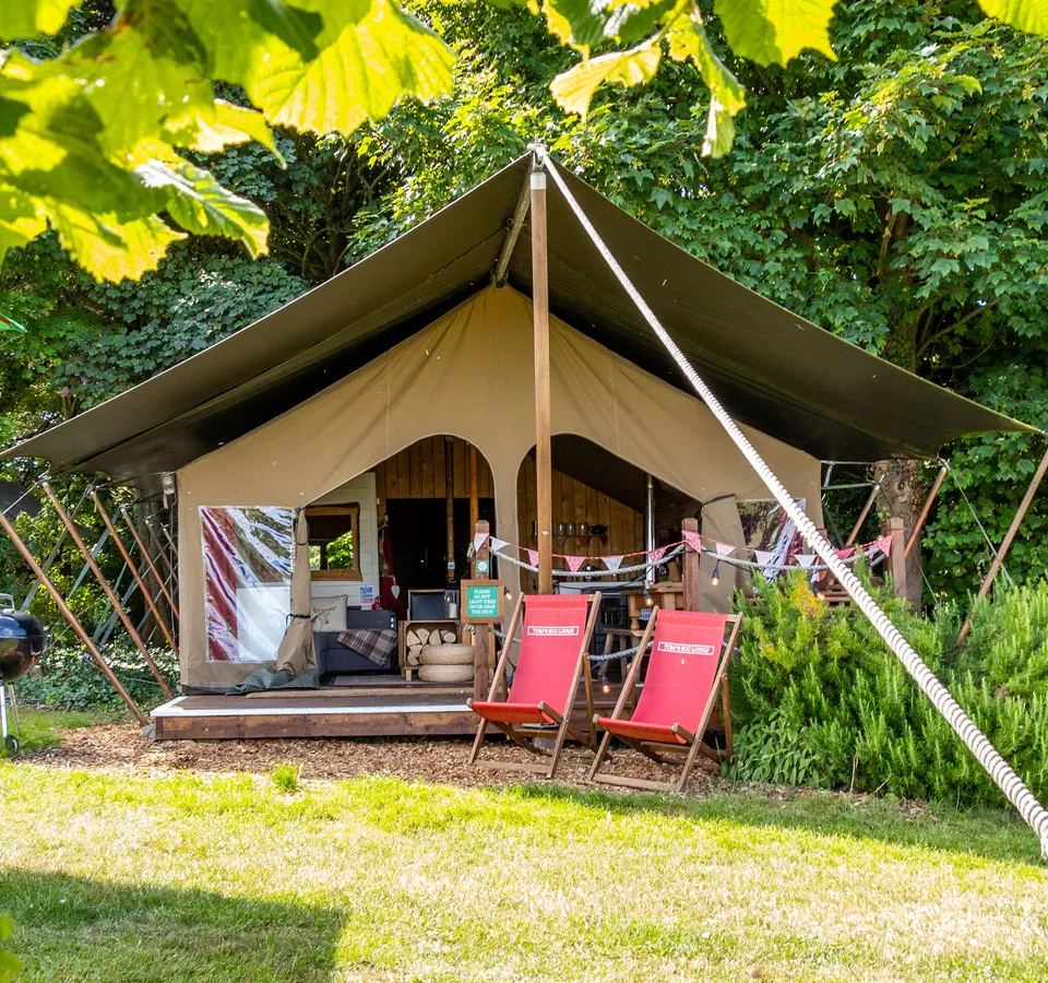 Safari-style eco lodge tent at Tapnell Farm on the Isle of Wight, with deck chairs and a barbecue in a leafy woodland setting