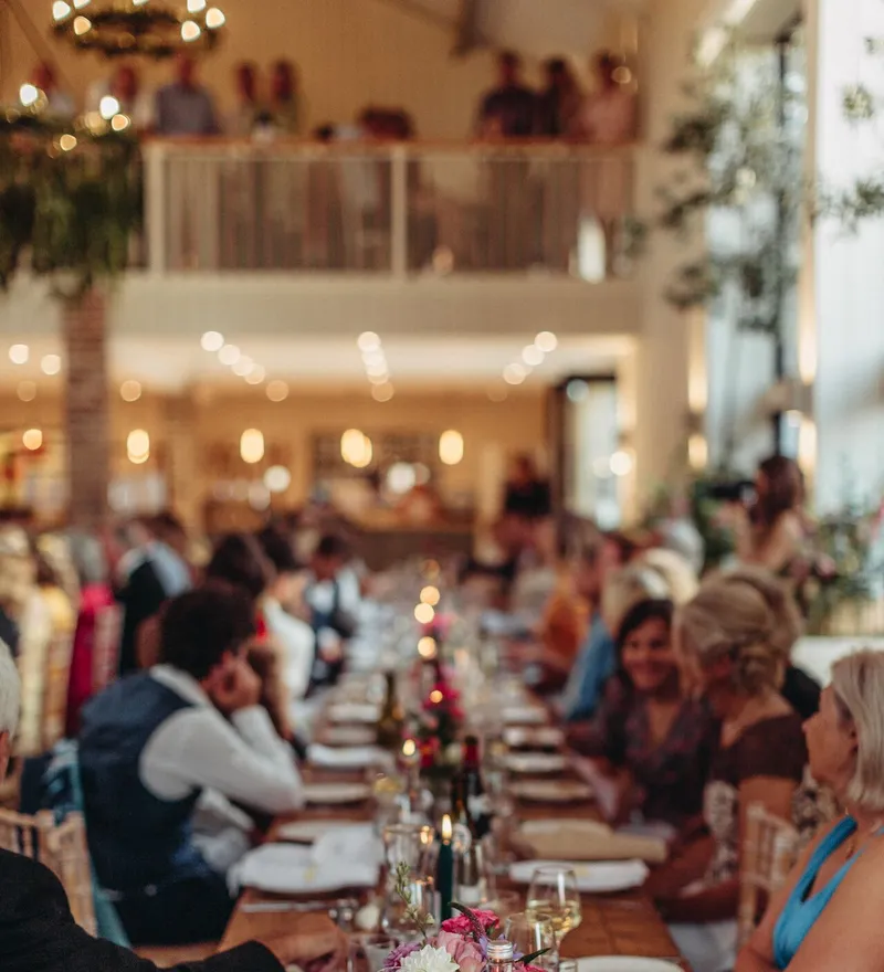 Guests seated at a long candlelit dining table during a wedding reception in a bright, airy venue