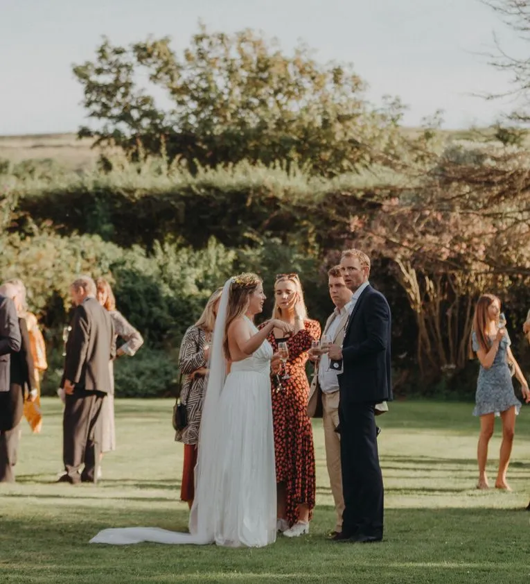 Wedding guests chatting and holding drinks on a lawn with the bride in the centre at an outdoor reception