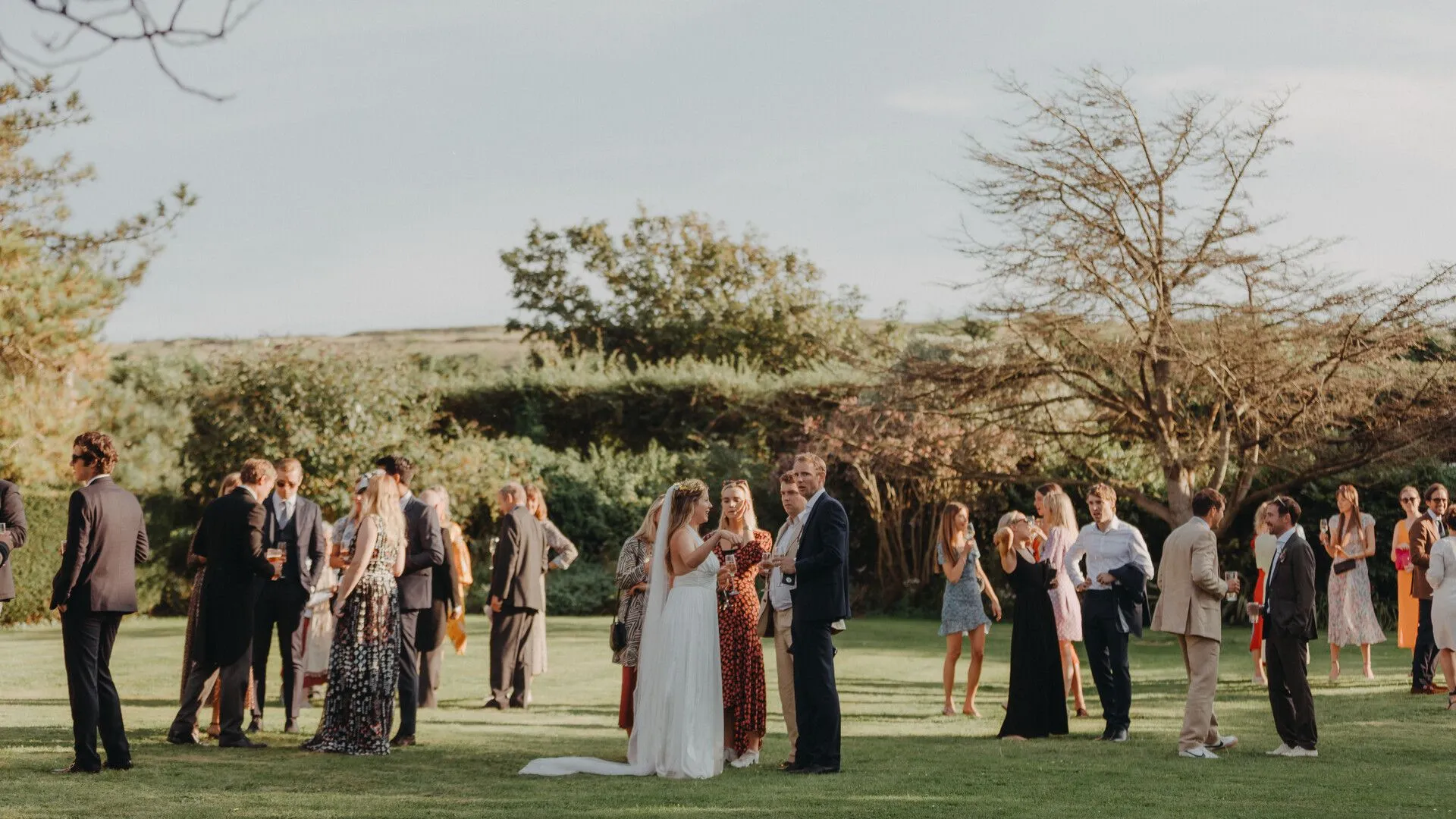 Wedding guests chatting and holding drinks on a lawn with the bride in the centre at an outdoor reception