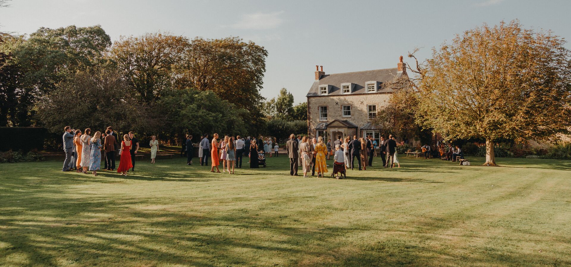 Wedding guests gathered on a lawn outside a country house in late afternoon light