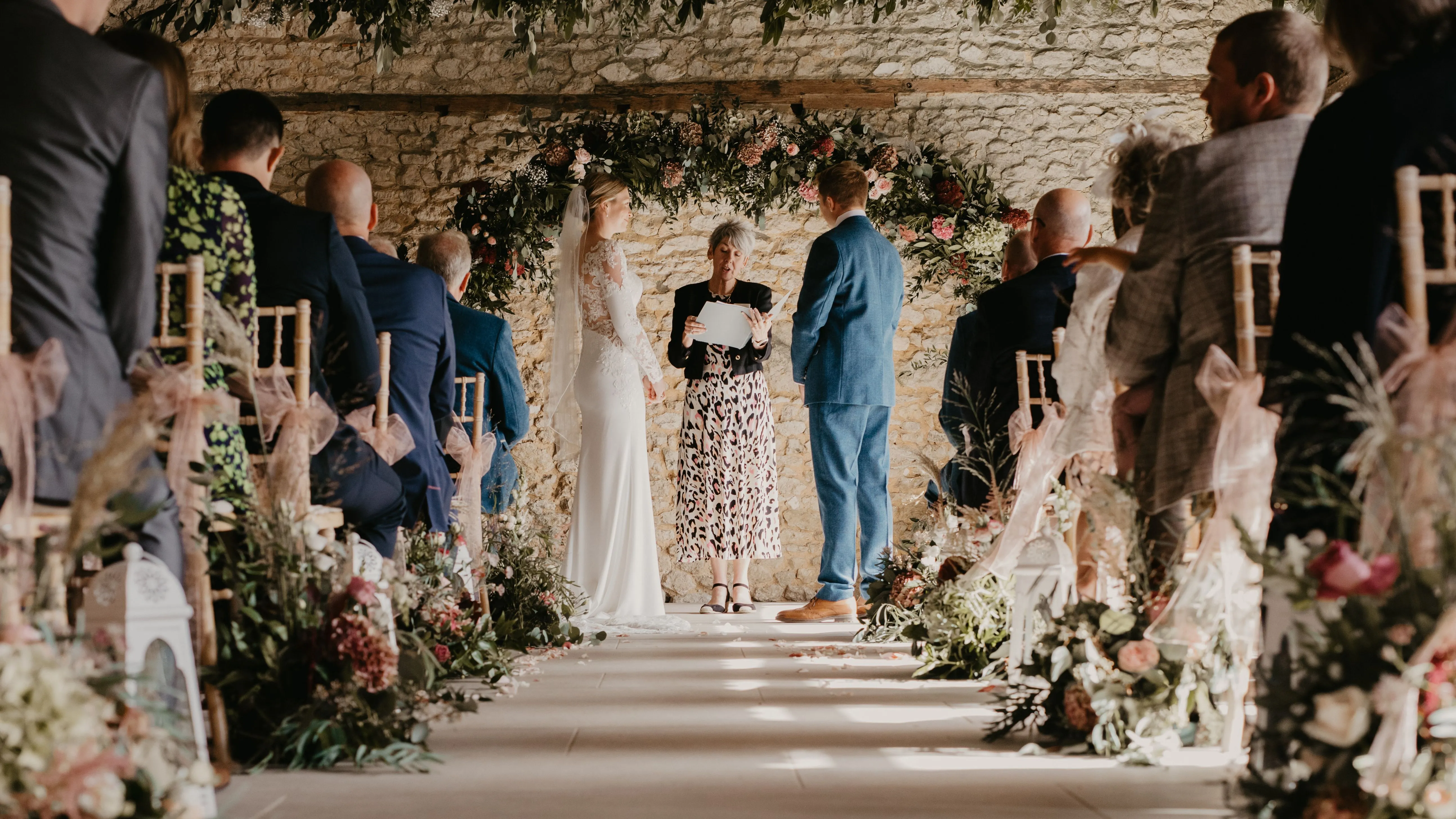 Bride and groom stand with officiant beneath a floral arch during an indoor barn wedding ceremony