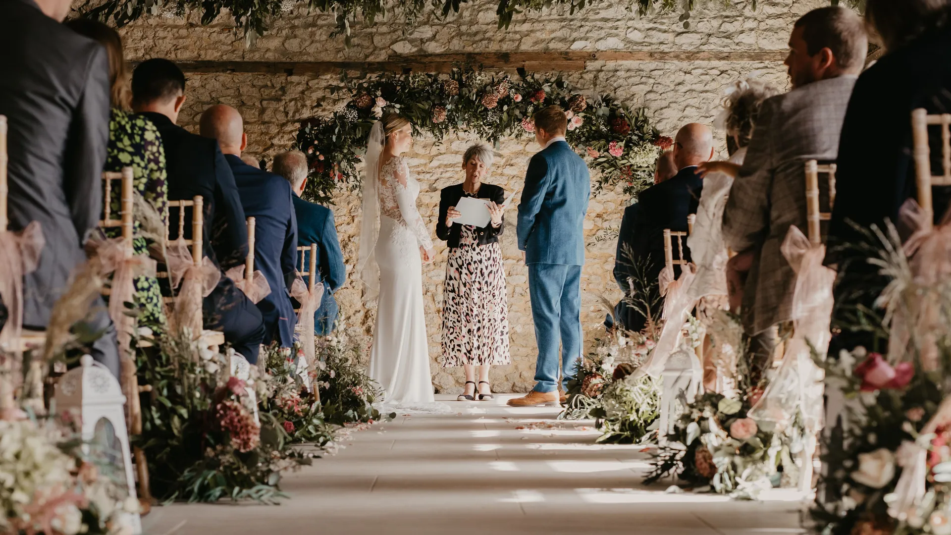 Bride and groom stand with officiant beneath a floral arch during an indoor barn wedding ceremony