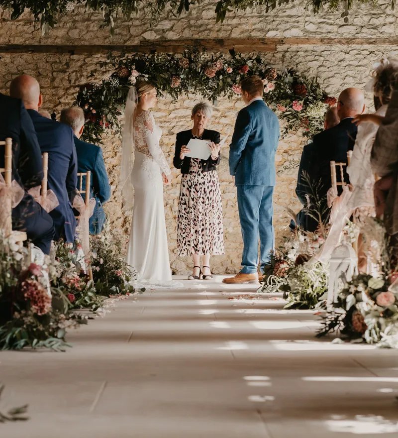 Bride and groom stand with officiant beneath a floral arch during an indoor barn wedding ceremony