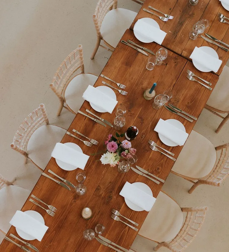 Overhead view of a long wooden dining table set with white plates, napkins, cutlery and glasses, with woven chairs and a floral centrepiece