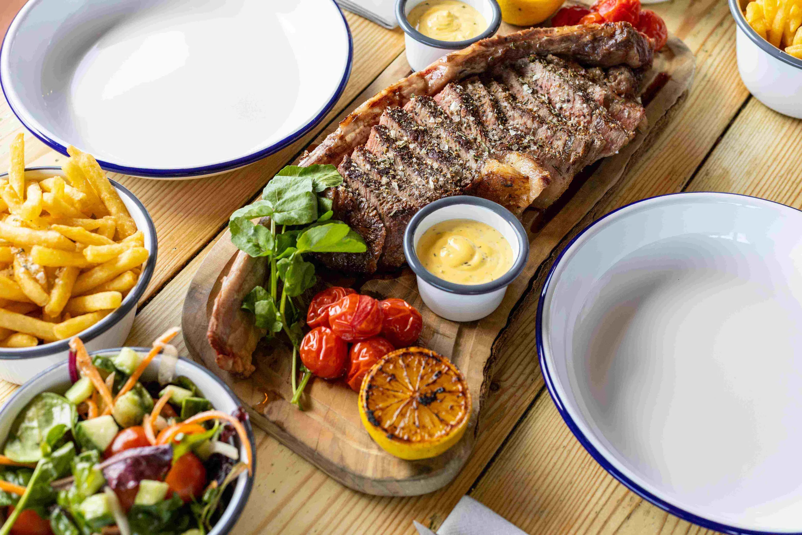 Grilled steak served with sauce, cherry tomatoes, salad, fries, and a charred lemon on a wooden platter.