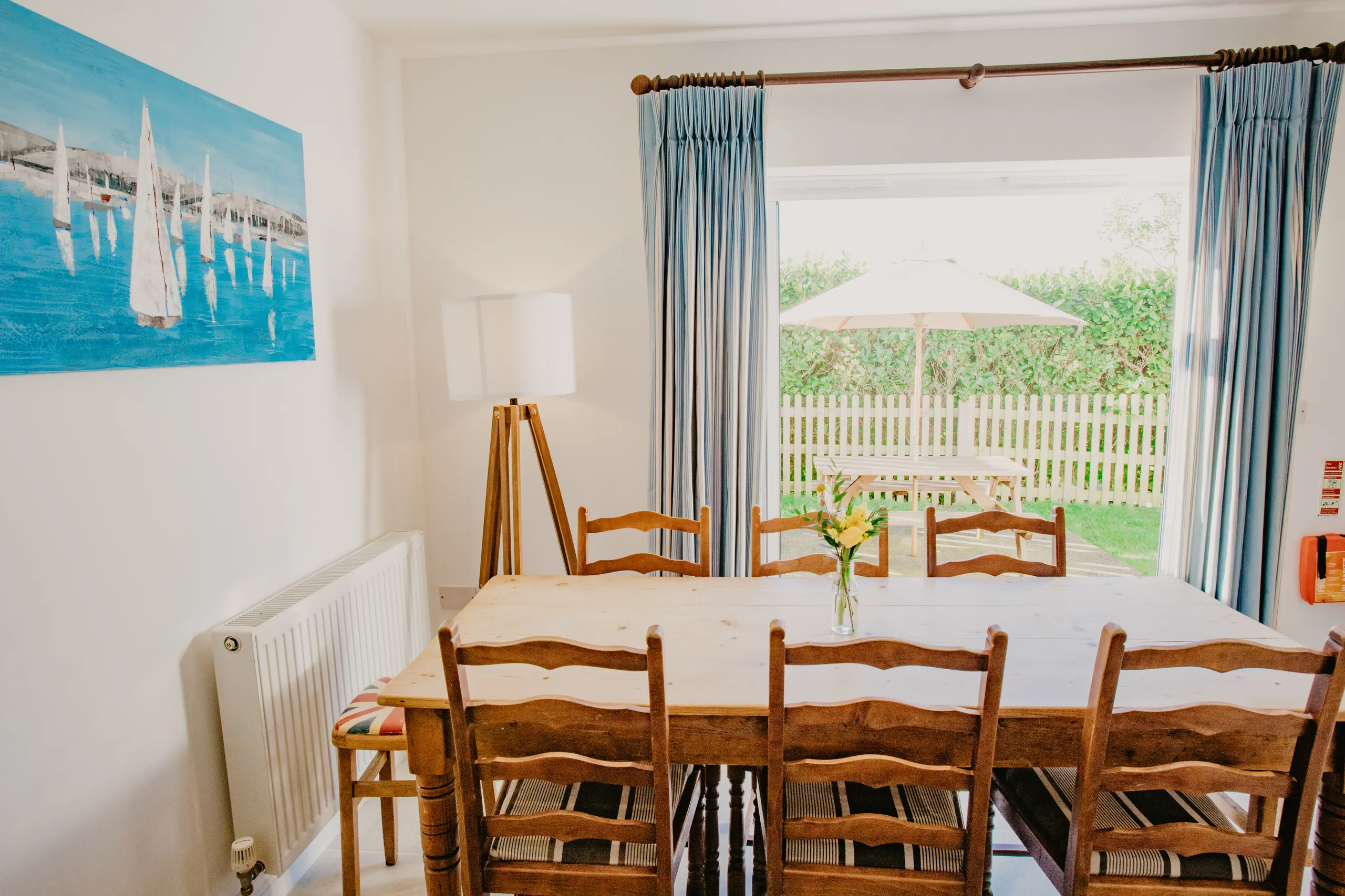 Dining table and chairs in a bright cottage dining room with blue curtains and garden patio beyond the sliding door