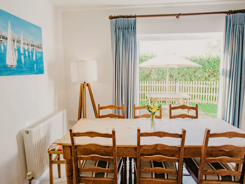 Dining table and chairs in a bright cottage dining room with blue curtains and garden patio beyond the sliding door