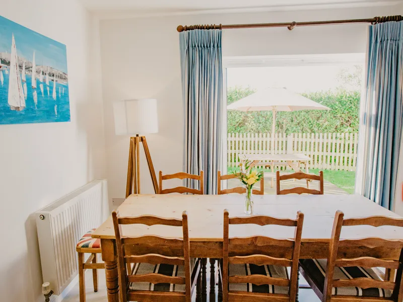 Dining table and chairs in a bright cottage dining room with blue curtains and garden patio beyond the sliding door
