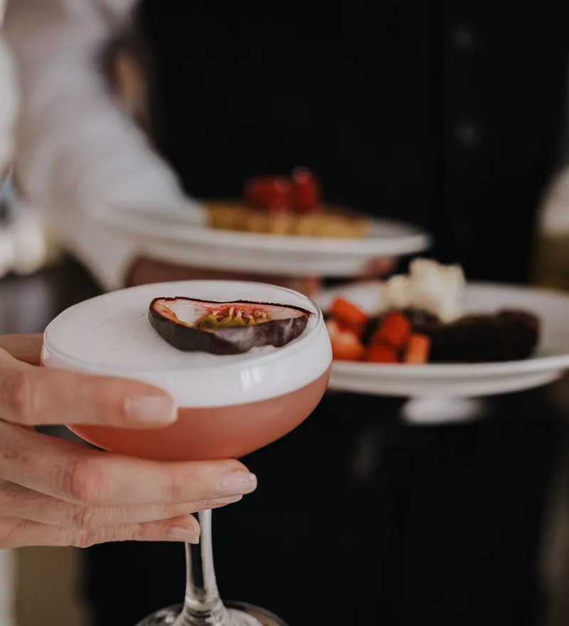 Close-up of a guest holding a pink cocktail with a foamy top and passion fruit garnish during a wedding reception