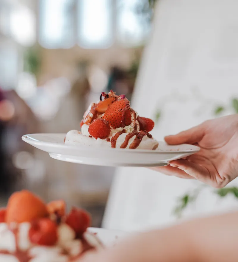 Hand holding a plate with a strawberry and cream dessert drizzled with chocolate sauce