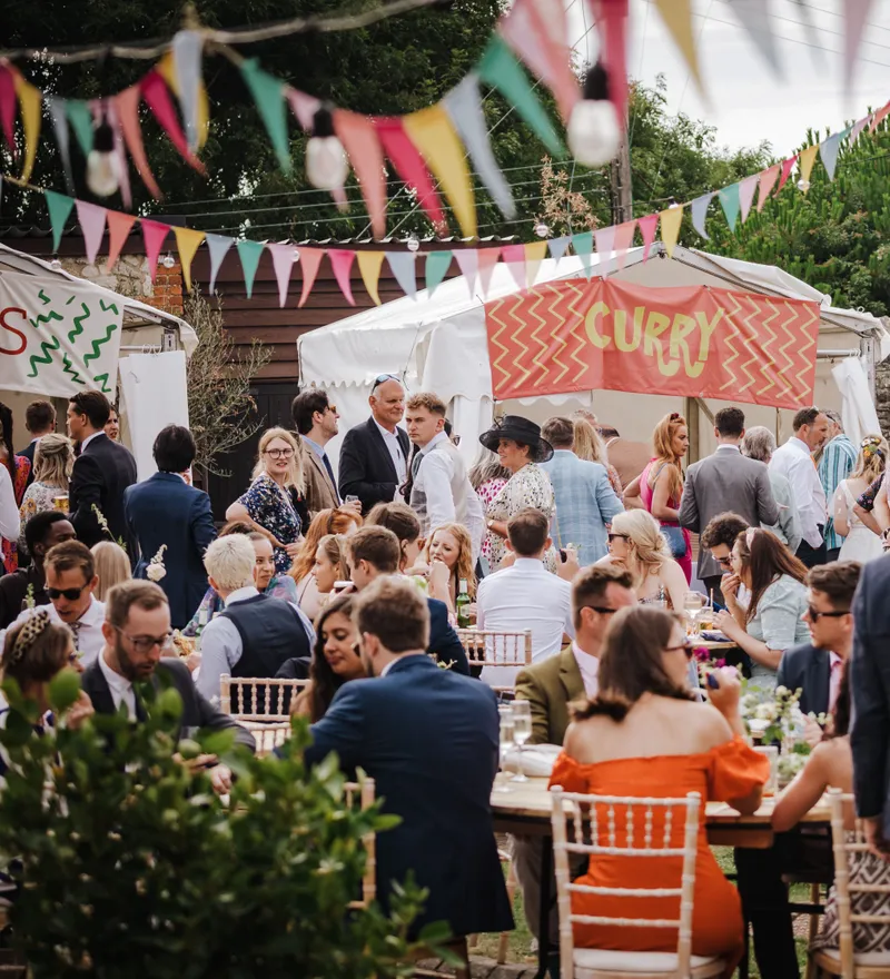 Wedding guests mingling outdoors beneath colourful bunting near a curry food stall