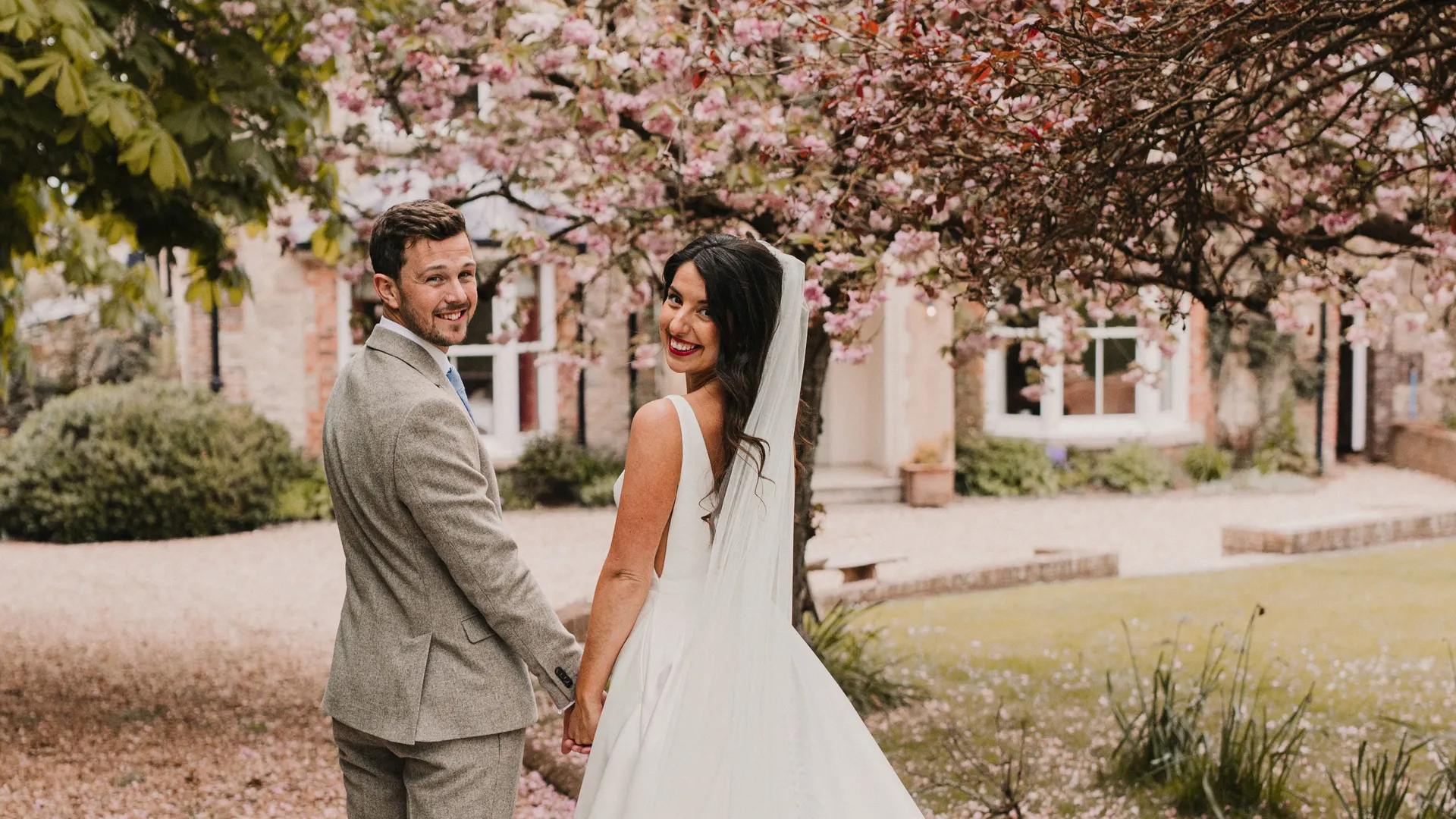 Bride and groom holding hands beneath pink blossom trees in a garden, looking back at the camera.