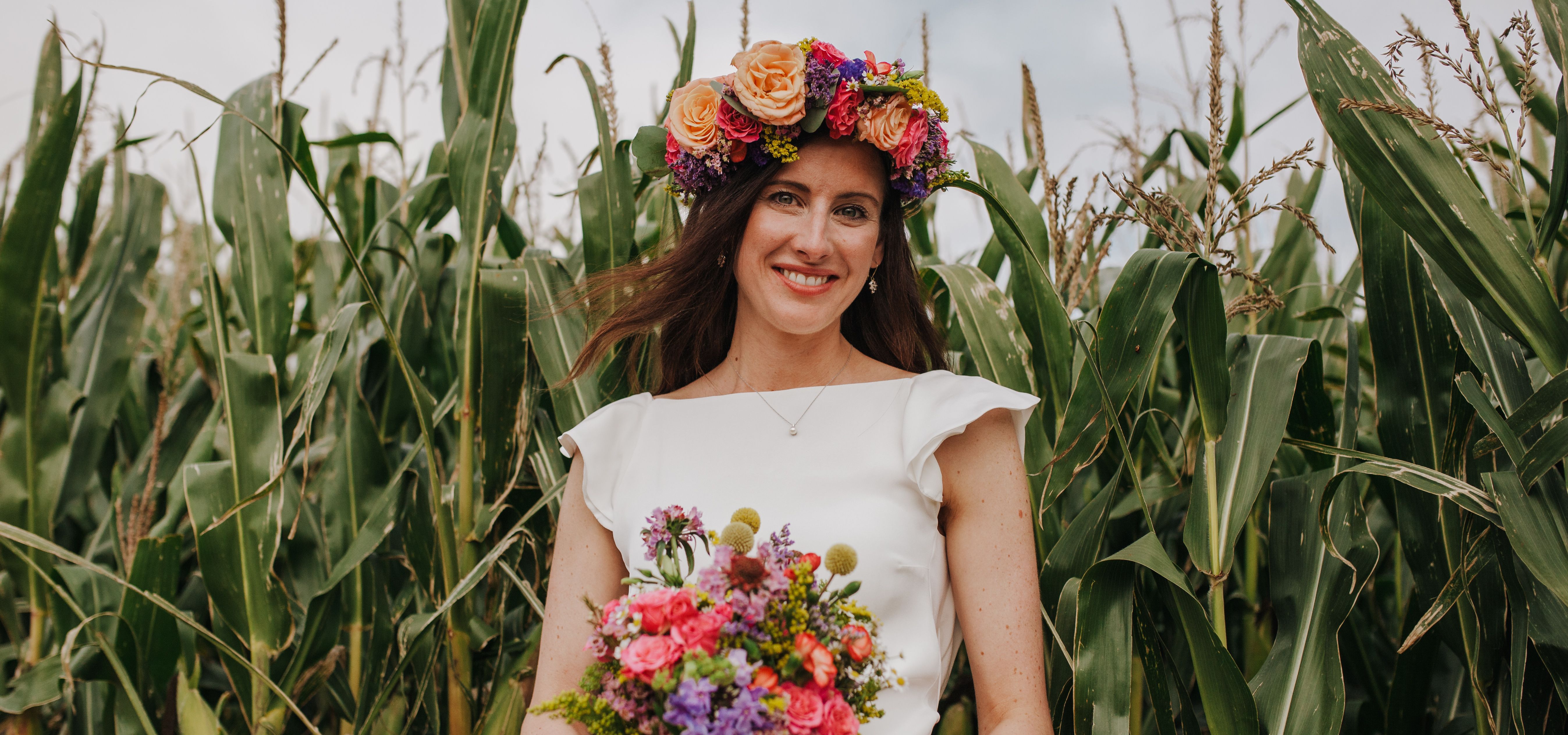 Bride in a white dress wearing a colourful flower crown and holding a bright bouquet in a cornfield
