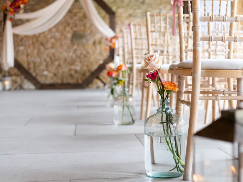 Glass bottle vases with roses and bright flowers lining the aisle beside wooden wedding chairs in a rustic barn venue