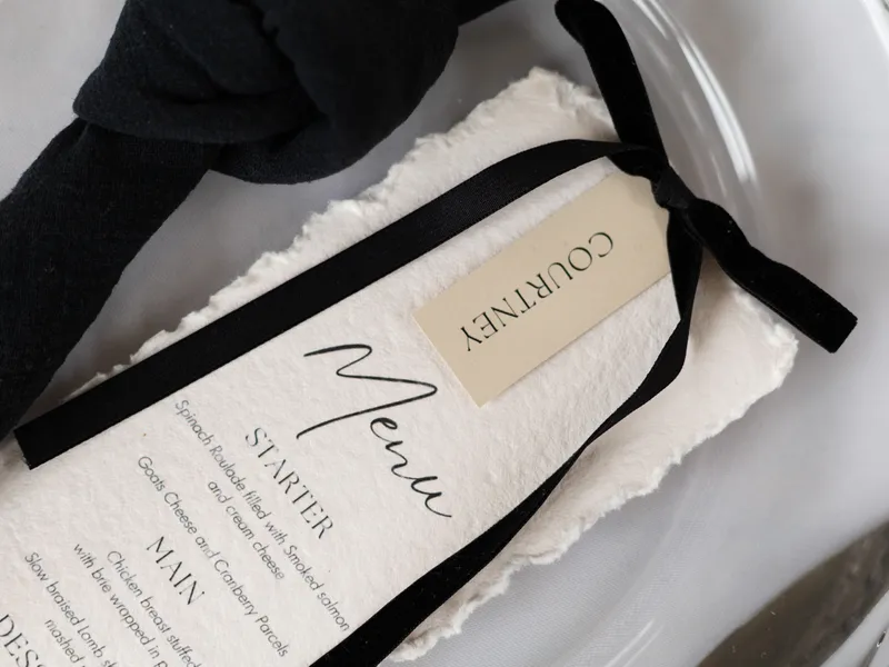 Wedding menu on textured paper tied with a black ribbon, beside white roller skates and a black bow accessory.