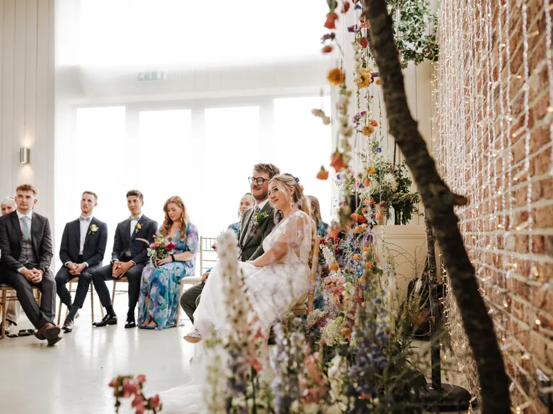 Bride and groom seated during wedding ceremony beside colourful floral installations and fairy lights