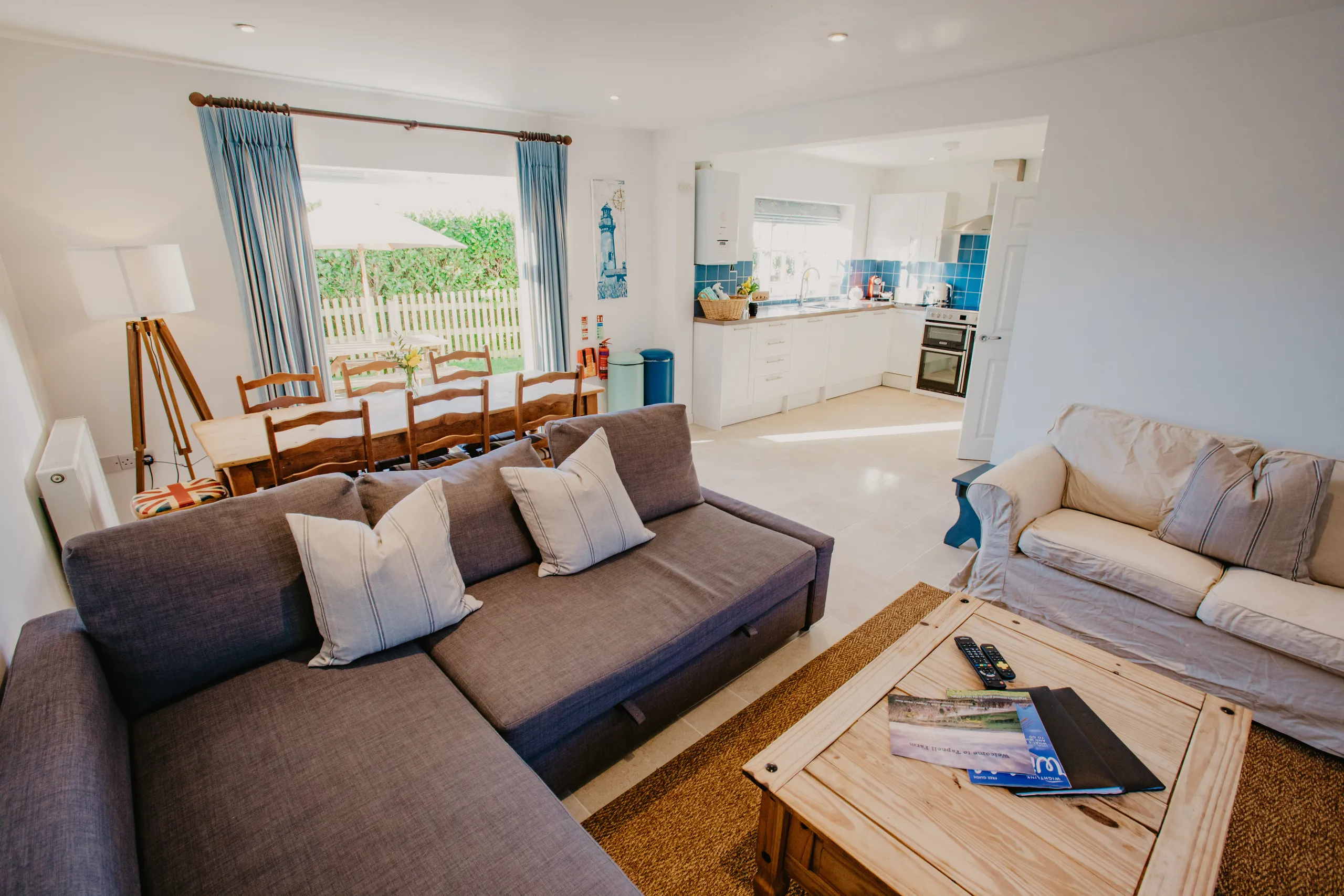 Open-plan cottage living and dining area with blue curtains, wooden dining table, sofas and a bright white kitchen with blue tiles