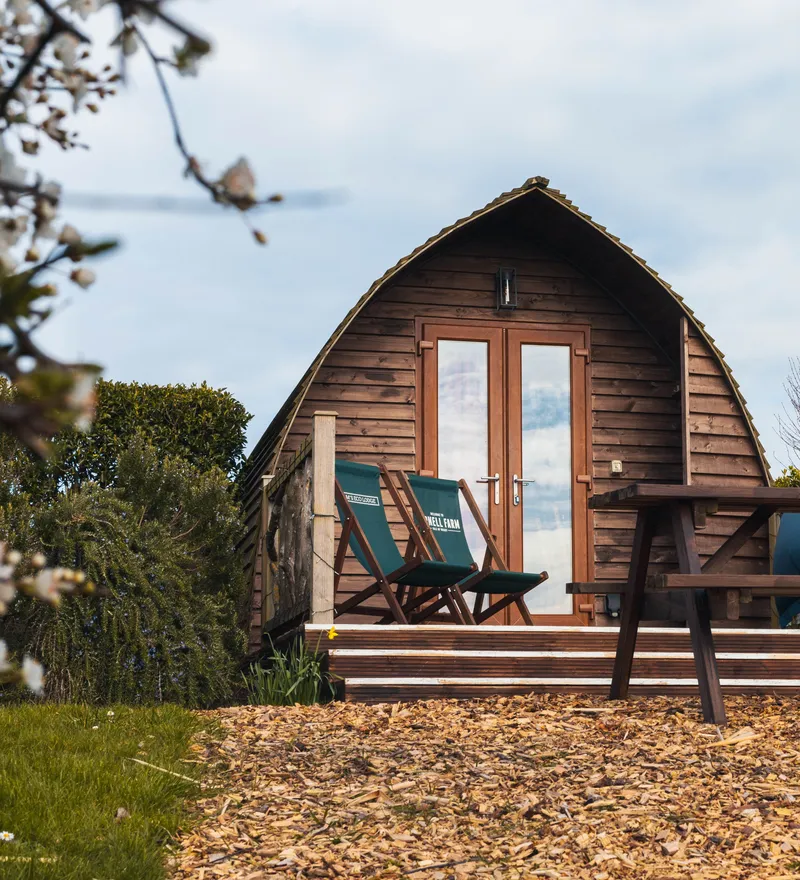 Wooden pod cabin with a small deck, two folding chairs and a picnic table in a garden setting
